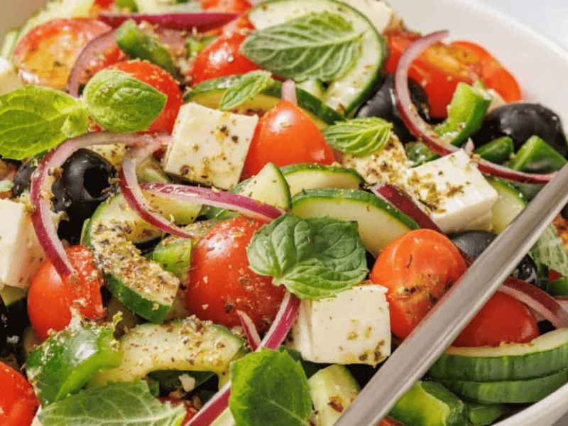 A bowl of Greek salad with cherry tomatoes, cucumber slices, red onion, black olives, feta cheese, and fresh mint leaves, sprinkled with herbs. A fork rests on the side.