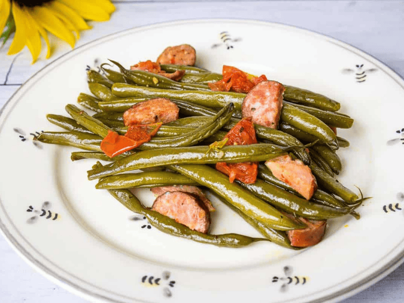 A plate of cooked Southern Green Beans with sliced sausages and tomato pieces on a white dish, placed near a sunflower.