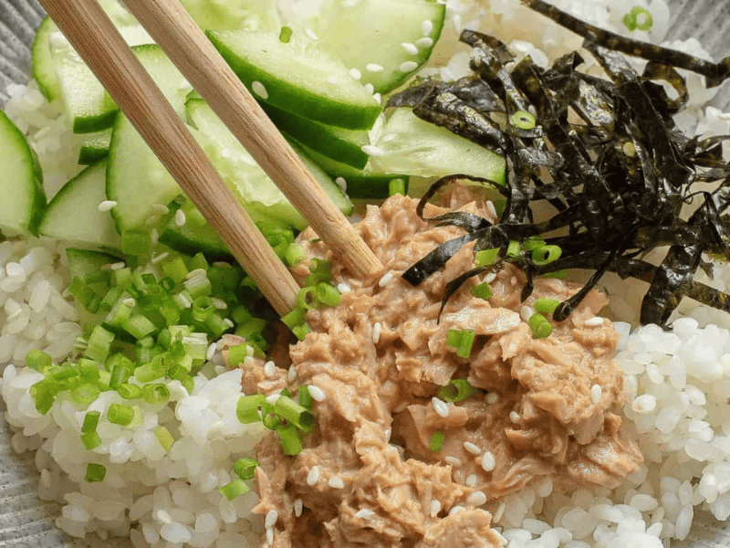 A bowl of white rice topped with sliced cucumber, shredded nori, chopped green onions, and a portion of tuna, with a pair of chopsticks resting on top.