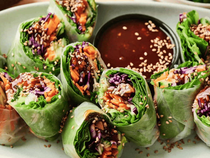 A plate of fresh vegetable spring rolls sliced in half, arranged around a dish of dipping sauce and sprinkled with sesame seeds.