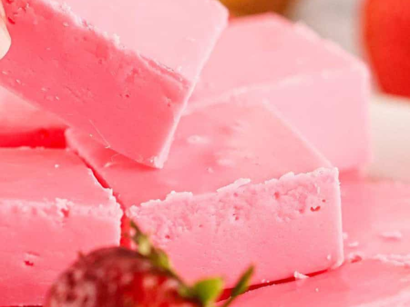 A hand picking up a square piece of pink fudge from a stack, with strawberries in the background.