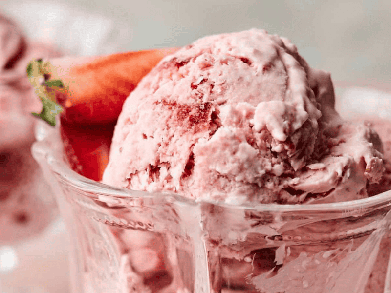 Close-up of scoops of strawberry ice cream in a glass dish, garnished with a fresh strawberry.