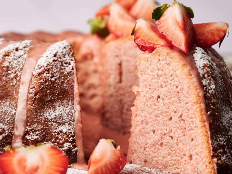 A sliced bundt cake with a pink interior, topped with powdered sugar, glaze, and fresh strawberries.