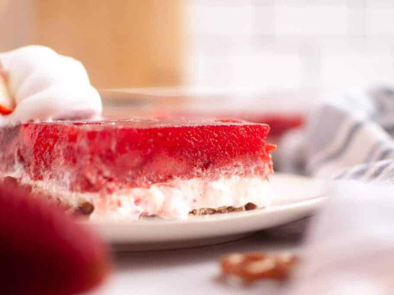 A slice of strawberry pretzel salad with whipped cream on a white plate, showing layers of red gelatin, creamy filling, and pretzel crust.