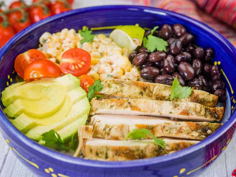 A blue bowl filled with street corn chicken rice bowl, featuring grilled chicken, corn, tomatoes, avocado, and black beans.