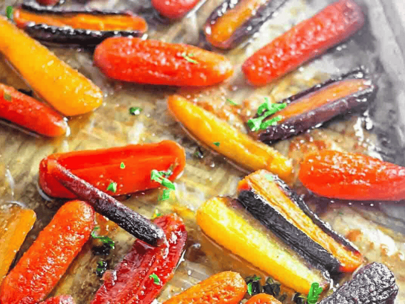 Assorted roasted carrots in orange, yellow, and purple colors arranged on a baking sheet with a light glaze and sprinkled with chopped herbs.
