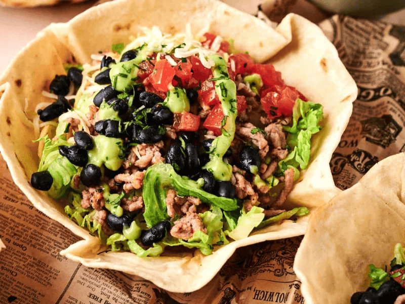 A taco salad served in an edible tortilla bowl, topped with ground meat, lettuce, black beans, diced tomatoes, avocado, and shredded cheese.