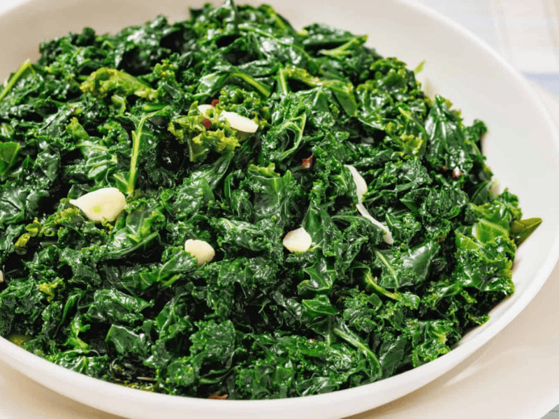A white bowl filled with sautéed kale and sliced garlic on a light-colored surface.