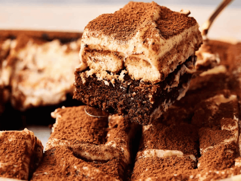 A close-up of a tray of tiramisu with a chocolate base, with one slice being lifted out, topped with dusted cocoa powder.