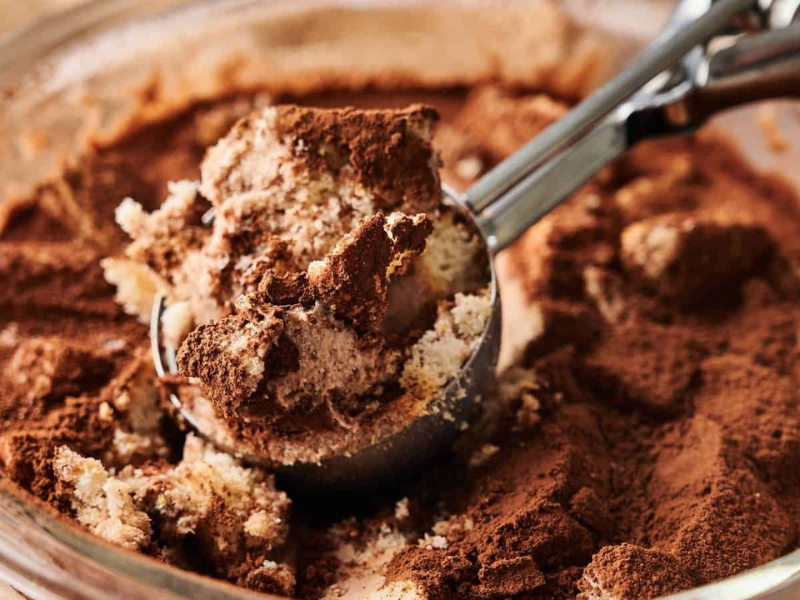 A close-up of a glass dish with tiramisu being scooped out, showing layers of cream, cake, and cocoa powder.
