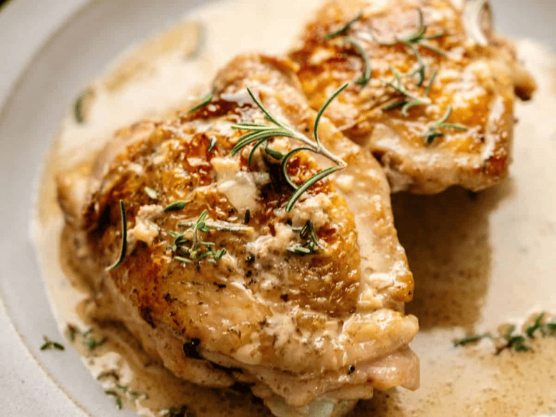Two pieces of cooked chicken with crispy skin are served on a plate, topped with creamy sauce and garnished with sprigs of fresh herbs.