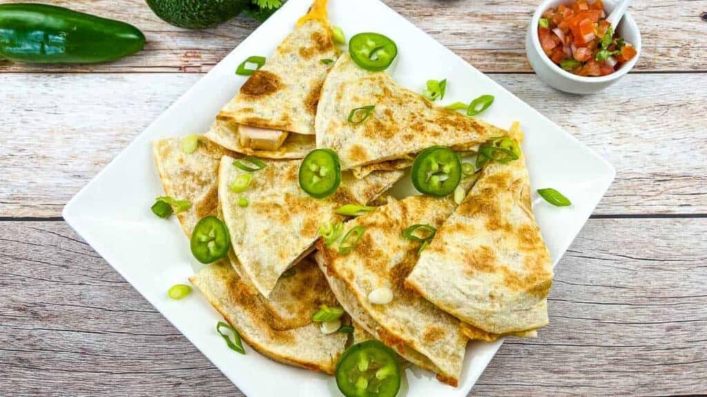 A plate of quesadilla wedges garnished with sliced jalapeños and chopped green onions, served on a white square dish with a small bowl of salsa on the side.