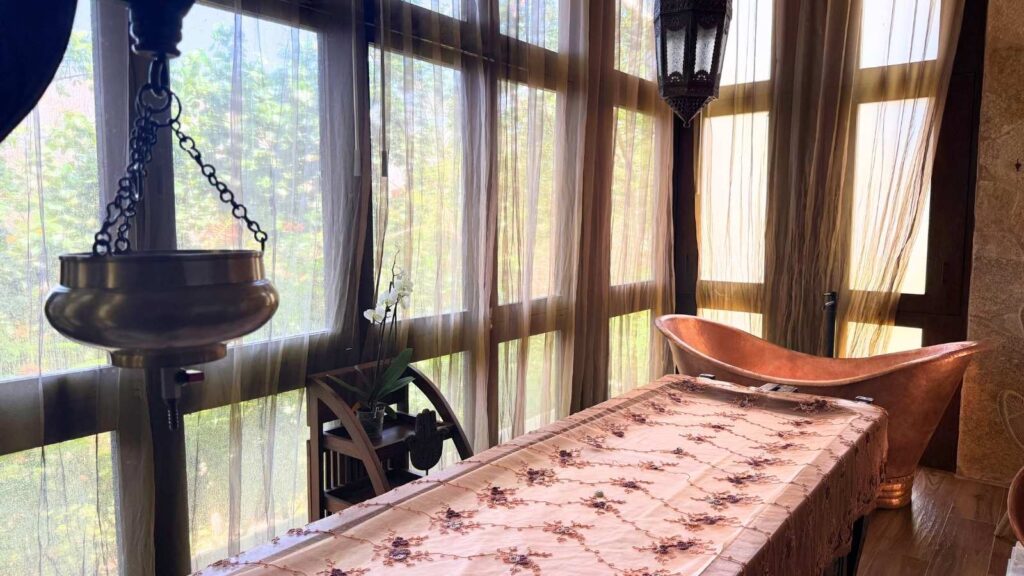 A spa room with sheer curtains, a decorative massage table, a copper bathtub, hanging lantern, and potted plant by tall windows evokes the tranquil ambiance often found at family wellness resorts.