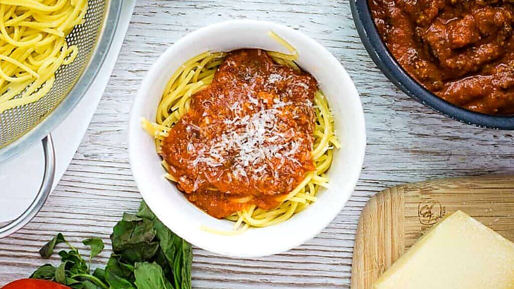 A bowl of spaghetti topped with red tomato sauce and grated cheese, placed on a white wooden surface near a block of cheese and fresh herbs—the rich sauce is perfectly blended using a Vitamix Immersion Blender for an irresistibly smooth texture.