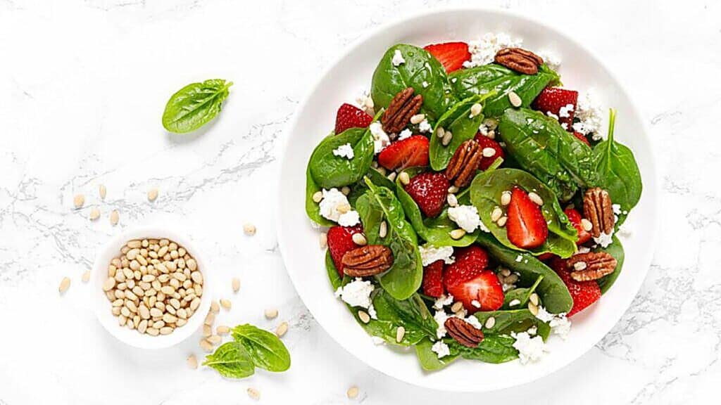 A white bowl contains spinach, strawberries, pecans, feta cheese, and pine nuts&mdash;a vibrant example of delicious maple syrup uses for enhancing salad dressings. Some spinach leaves and a small bowl of pine nuts are on the marble surface beside it.