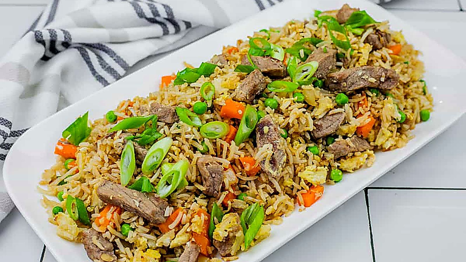 Steak fried rice on a white platter on white tiles with striped cloth on the side.