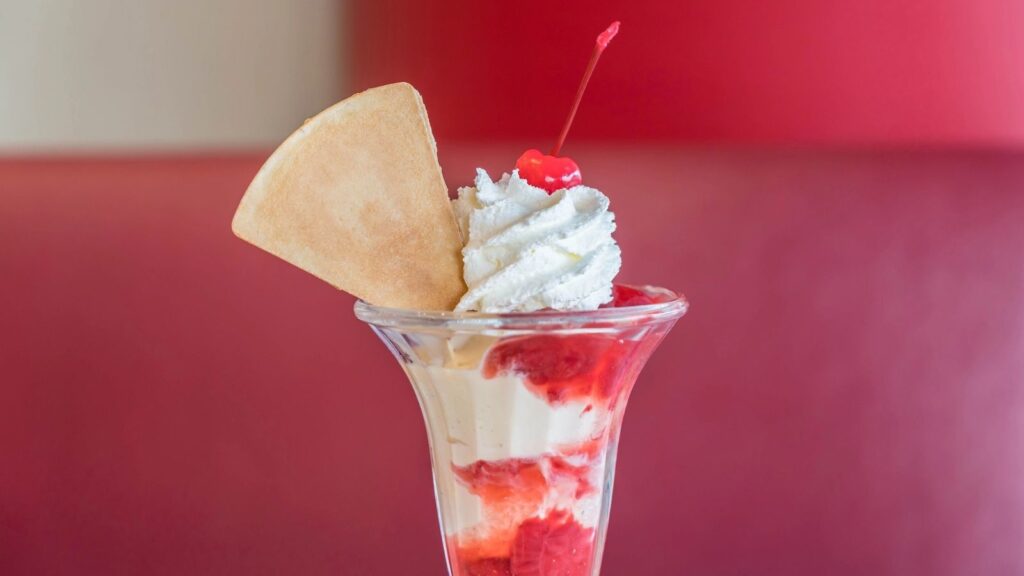 A glass sundae with vanilla ice cream, strawberry sauce, whipped cream, a cherry on top, and a wafer cookie, set against a red background.