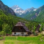 Two wooden houses with fenced yards sit in a green valley surrounded by forested mountains under a clear blue sky, with a dirt path curving past the homes—a tranquil scene that captures the allure of Slovenia travel.