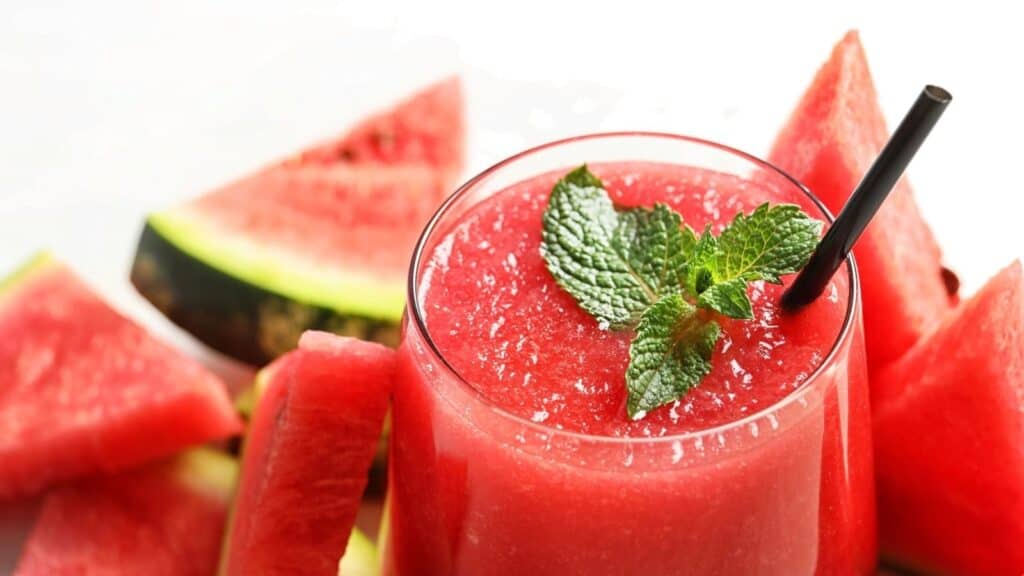 A glass of watermelon smoothie with a black straw and mint garnish, surrounded by fresh watermelon slices.