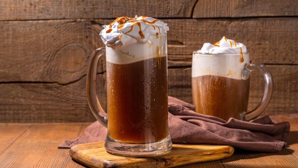 Two glass mugs filled with dark brown root beer topped with whipped cream and caramel drizzle sit on a wooden table with a brown cloth and wooden background.