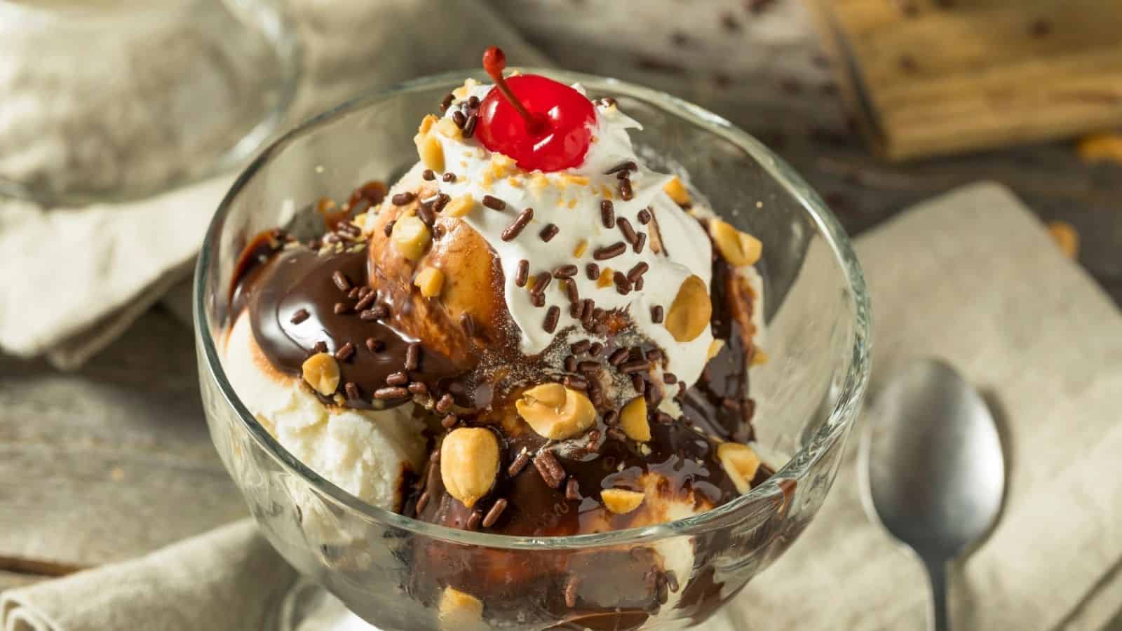 A glass bowl of ice cream topped with chocolate sauce, whipped cream, peanuts, chocolate sprinkles, and a cherry, with a spoon beside it on a wooden table.