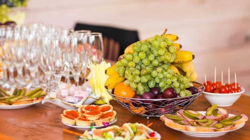 A table set with champagne glasses, a fruit basket with bananas and grapes, a bowl of cherry tomatoes, and assorted appetizers on plates creates a festive spread that&rsquo;s perfect for gatherings where potluck rules invite everyone to share their favorite dishes.