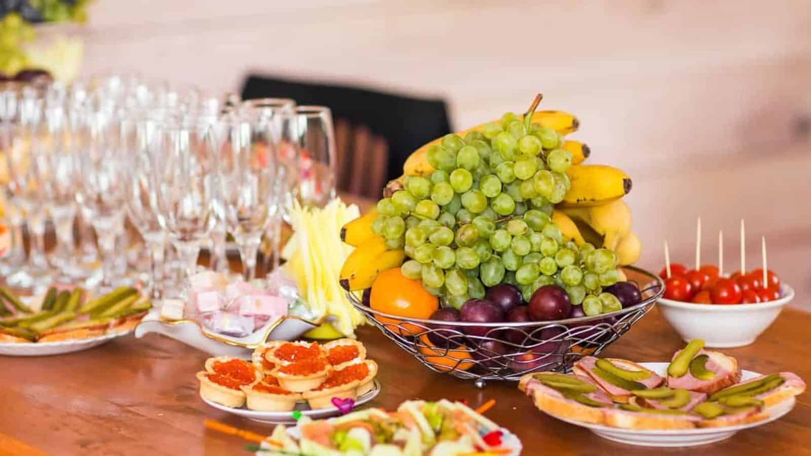 A table set with champagne glasses, a fruit basket with bananas and grapes, a bowl of cherry tomatoes, and assorted appetizers on plates creates a festive spread that&rsquo;s perfect for gatherings where potluck rules invite everyone to share their favorite dishes.