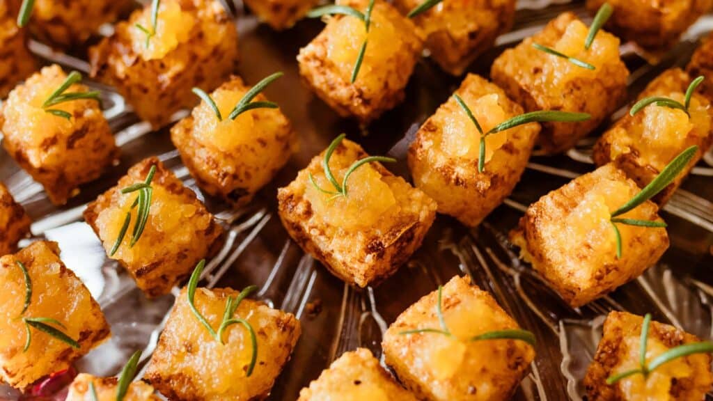 Small square appetizers topped with a yellow spread and garnished with sprigs of rosemary, arranged neatly on a glass platter—perfect bites to celebrate World Tofu Day.