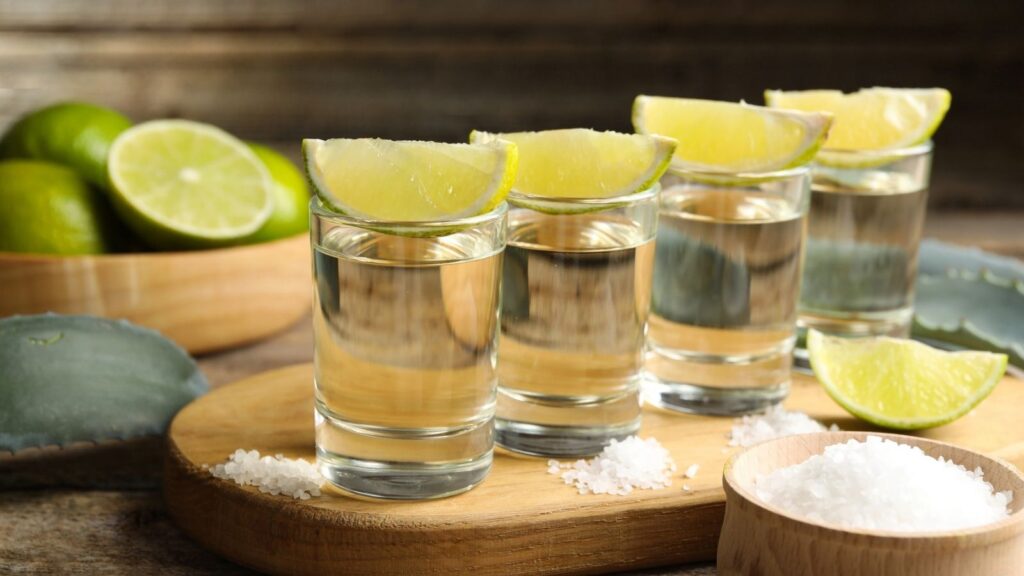 Four tequila shots with lime wedges on a wooden board, surrounded by salt piles and sliced limes in the background—perfect for celebrating National Tequila Day in style.