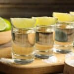 Four tequila shots with lime wedges on a wooden board, surrounded by salt piles and sliced limes in the background—perfect for celebrating National Tequila Day in style.