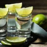 Three tequila shots with lime wedges on top, accompanied by lime slices, an agave leaf, ice cubes, and a small pile of salt on a slate tray with a wooden background—an inviting scene that hints at both the tradition and variety reflected in today’s tequila prices.
