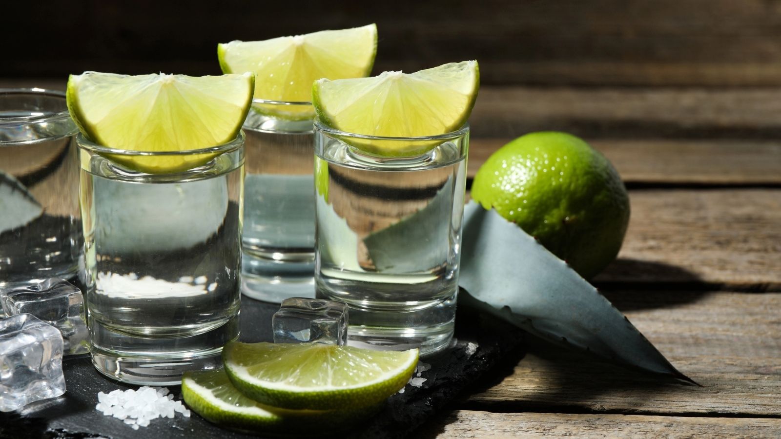 Three tequila shots with lime wedges on top, accompanied by lime slices, an agave leaf, ice cubes, and a small pile of salt on a slate tray with a wooden background—an inviting scene that hints at both the tradition and variety reflected in today’s tequila prices.