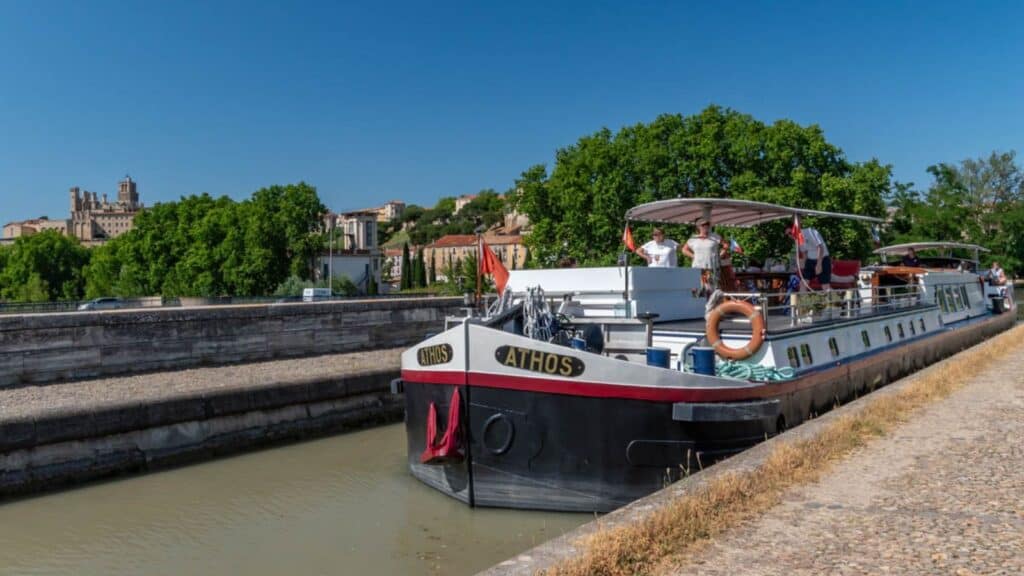 A canal barge named "Athos" is docked along a stone pathway, with people on board enjoying a southern France barge cruise and a picturesque town with historic buildings in the background.
