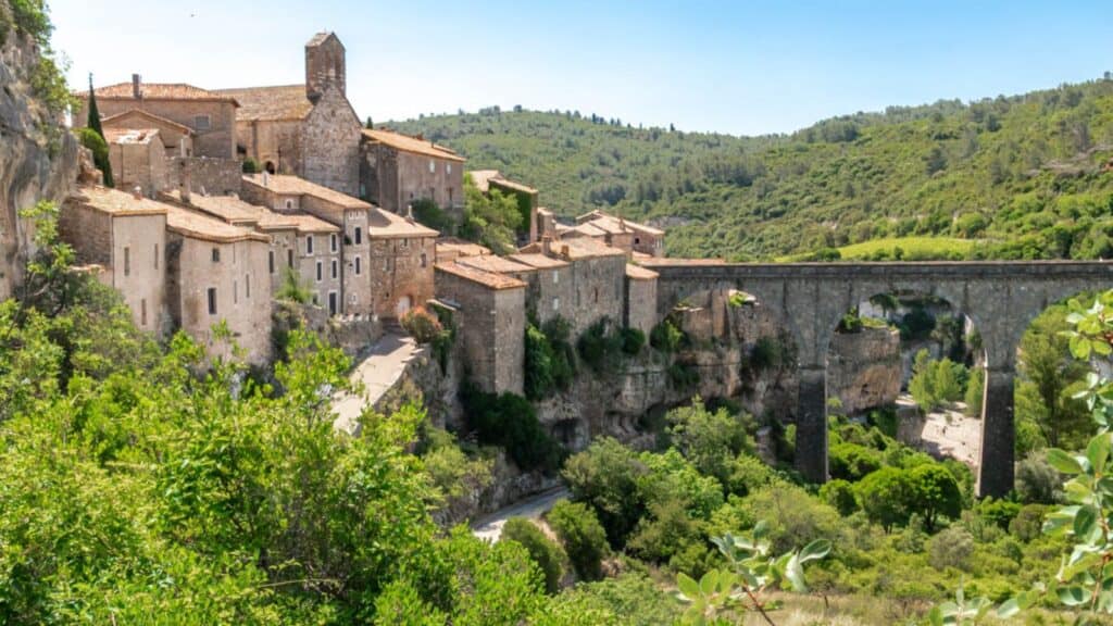 A historic stone village with terracotta roofs sits on a hill beside an arched stone bridge, surrounded by green trees and rolling hills under a clear blue sky—a picturesque scene often admired during a southern France barge cruise.