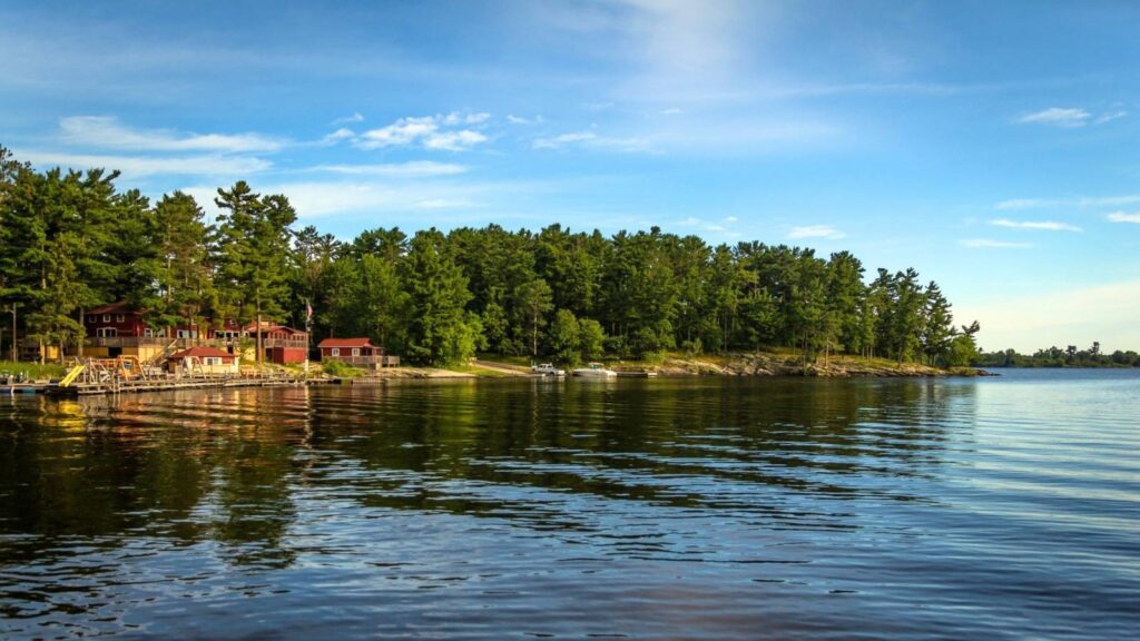A calm lake with reflections of trees and a clear sky, with several red cabins and boats along the forested shoreline.