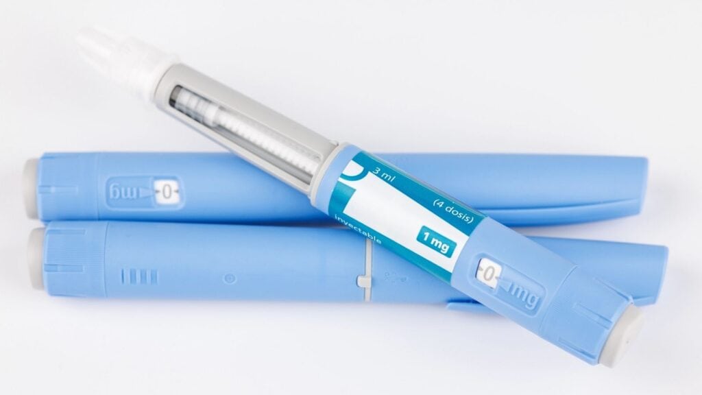 Three blue insulin-style injection pens&mdash;one with a clear window showing the dose and a label reading "1 mg"&mdash;are displayed on a white surface, highlighting the GLP-1 drugs that are increasingly discussed in restaurants and public spaces for their role in health management.