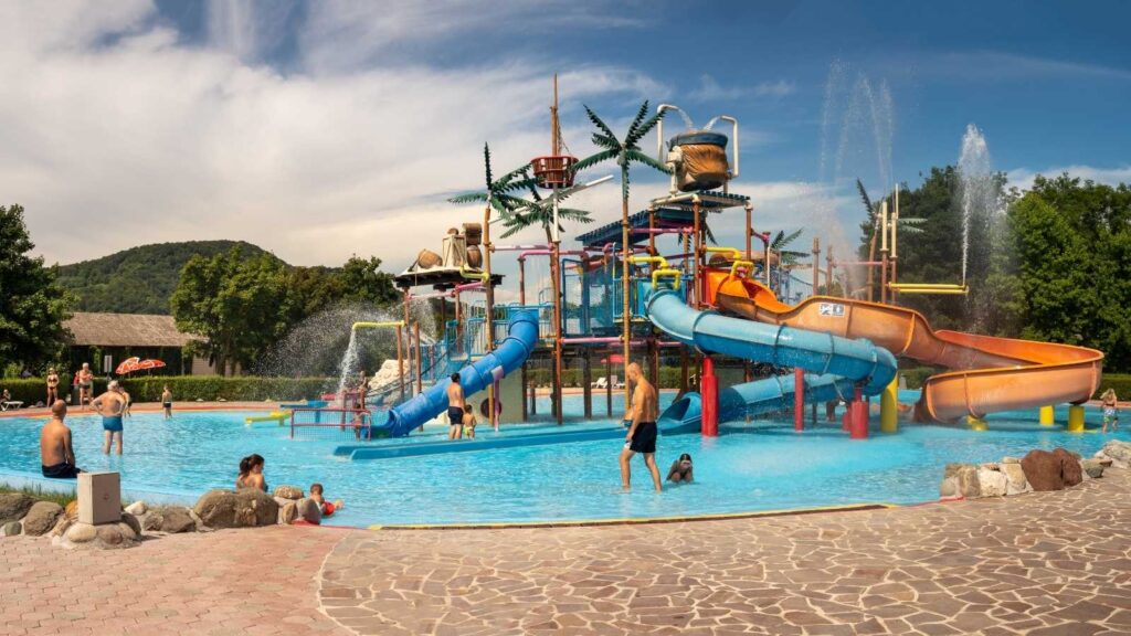 Americans discover Slovenia as they enjoy a vibrant water park filled with slides, splash features, and shallow pools on a sunny day. Trees and hills provide a picturesque backdrop, capturing the country&rsquo;s natural beauty.