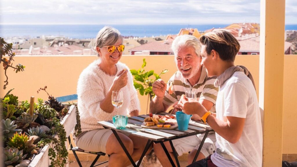 Three people sit at an outdoor table on a sunny terrace, sharing food and drinks in true blue zones fashion, with buildings and the sea visible in the background.