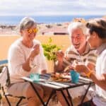 Three people sit at an outdoor table on a sunny terrace, sharing food and drinks in true blue zones fashion, with buildings and the sea visible in the background.