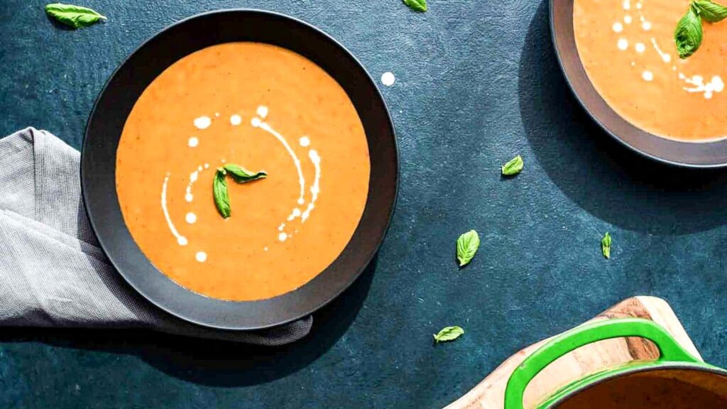 Two bowls of creamy tomato soup garnished with basil leaves, with a gray napkin and scattered basil on a dark background.