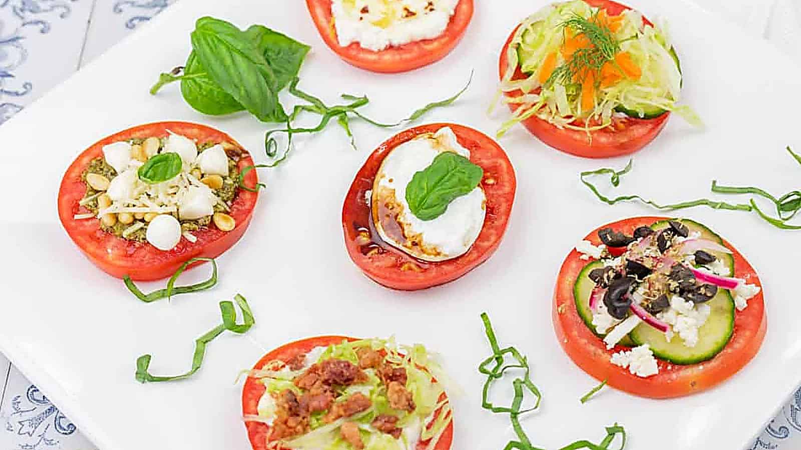 Six tomato slices topped with various ingredients, including cheese, lettuce, olives, herbs, and garnishes, arranged on a white plate with basil leaves and shredded greens.