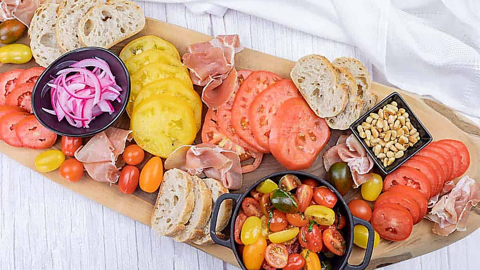 A wooden board with sliced bread, red and yellow tomato slices, cured meat, pickled onions, pine nuts, and a bowl of mixed cherry tomatoes.