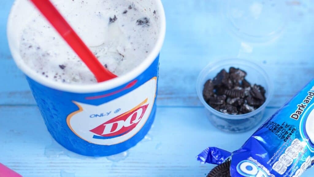 A Dairy Queen cup with an Oreo Blizzard, a red spoon, a small cup of crushed Oreos, and a pack of Oreo cookies on a blue wooden surface.