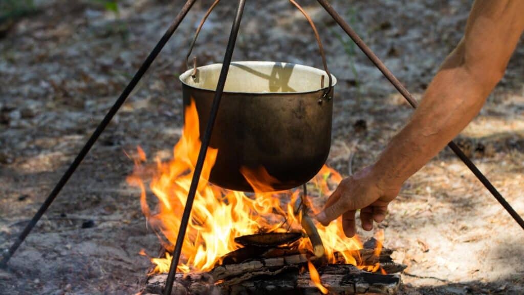 A metal pot hangs over an open fire on a tripod stand outdoors as a person adjusts the firewood beneath it.