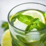 A glass of iced lime and mint drink, reminiscent of a classic mojito cocktail, with a green striped straw, garnished with fresh mint leaves and lime slices.