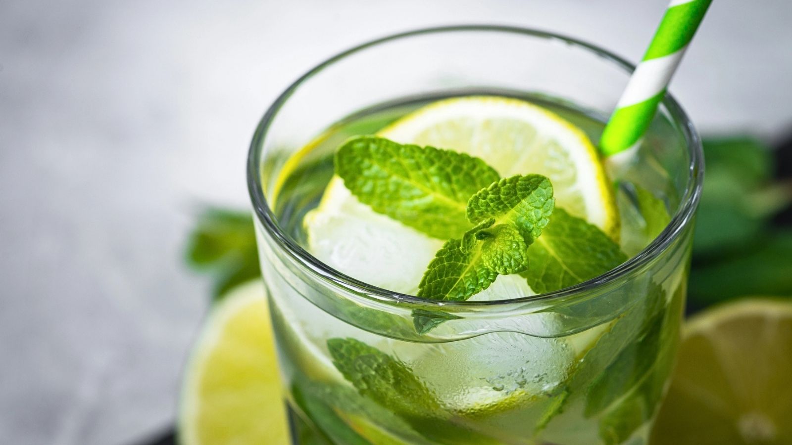 A glass of iced lime and mint drink, reminiscent of a classic mojito cocktail, with a green striped straw, garnished with fresh mint leaves and lime slices.