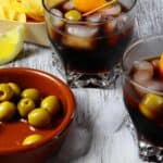 Two glasses of iced dark vermouth garnished with orange peel, a bowl of green olives, a wedge of lemon, and tortilla chips rest on a rustic wooden surface.