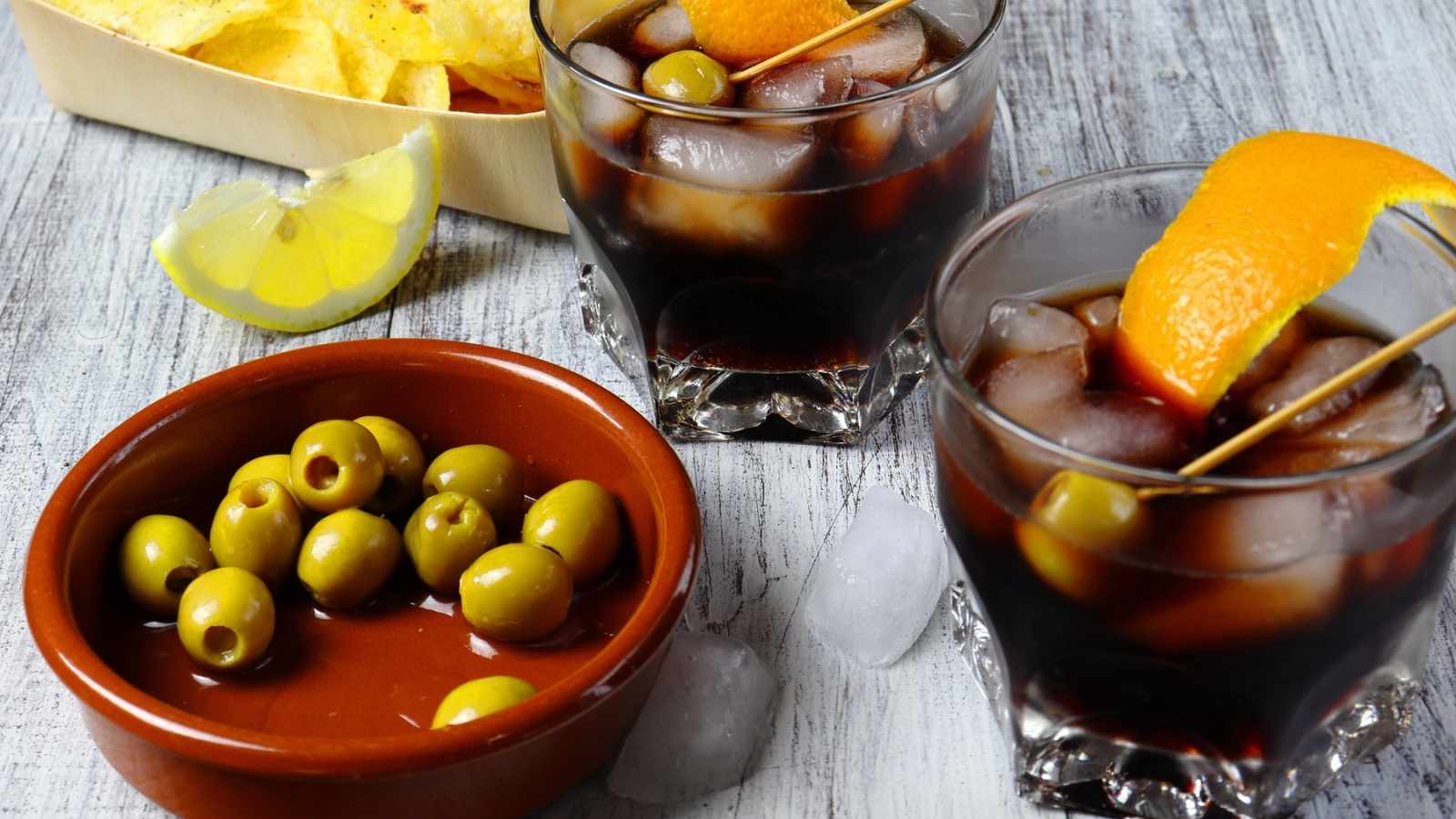 Two glasses of iced dark vermouth garnished with orange peel, a bowl of green olives, a wedge of lemon, and tortilla chips rest on a rustic wooden surface.
