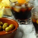 Two glasses of dark iced vermouth drinks with orange peels, a bowl of green olives, a lemon wedge, and a tray of potato chips rest on a wooden surface with ice cubes nearby.