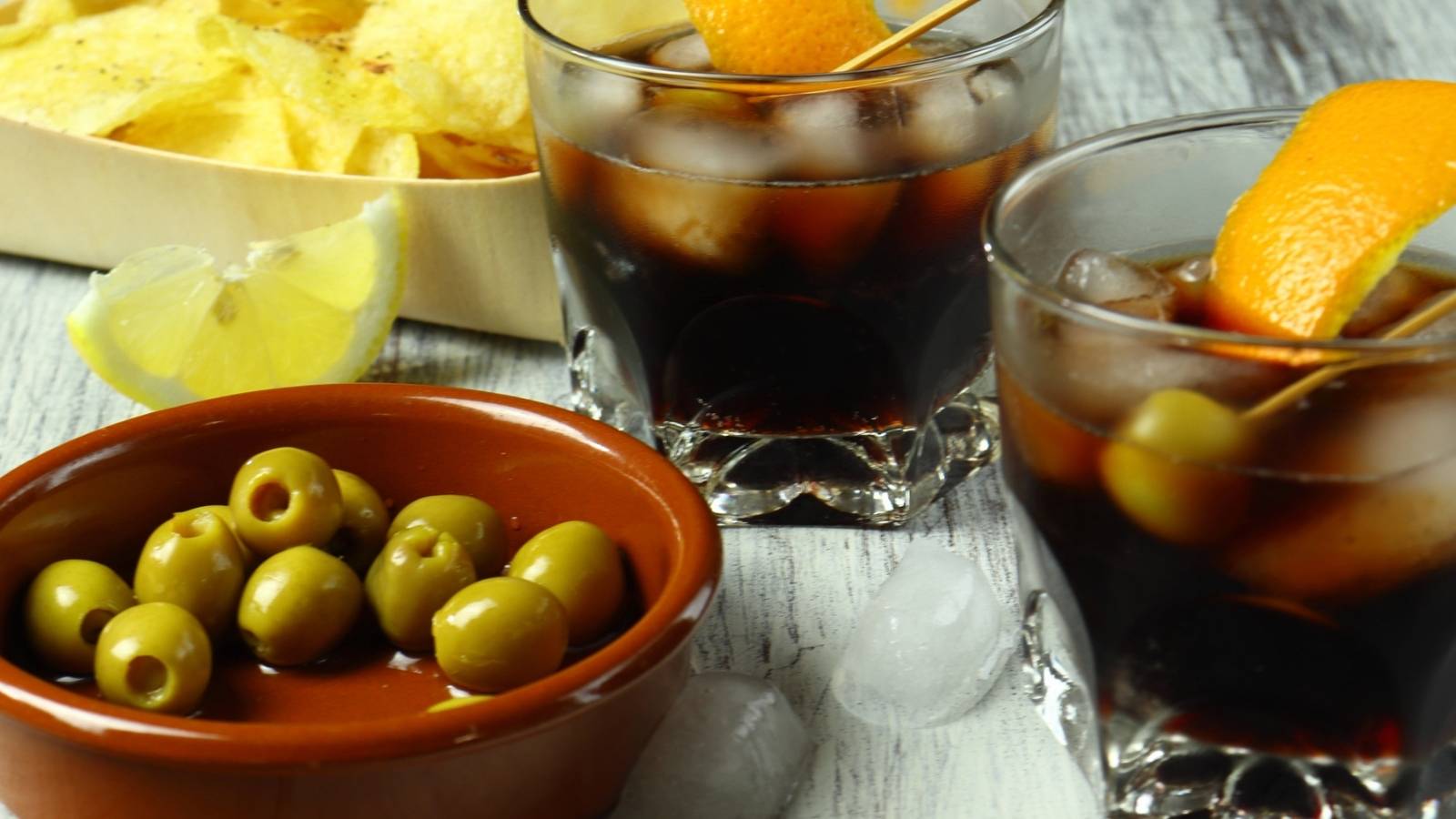 Two glasses of dark iced vermouth drinks with orange peels, a bowl of green olives, a lemon wedge, and a tray of potato chips rest on a wooden surface with ice cubes nearby.